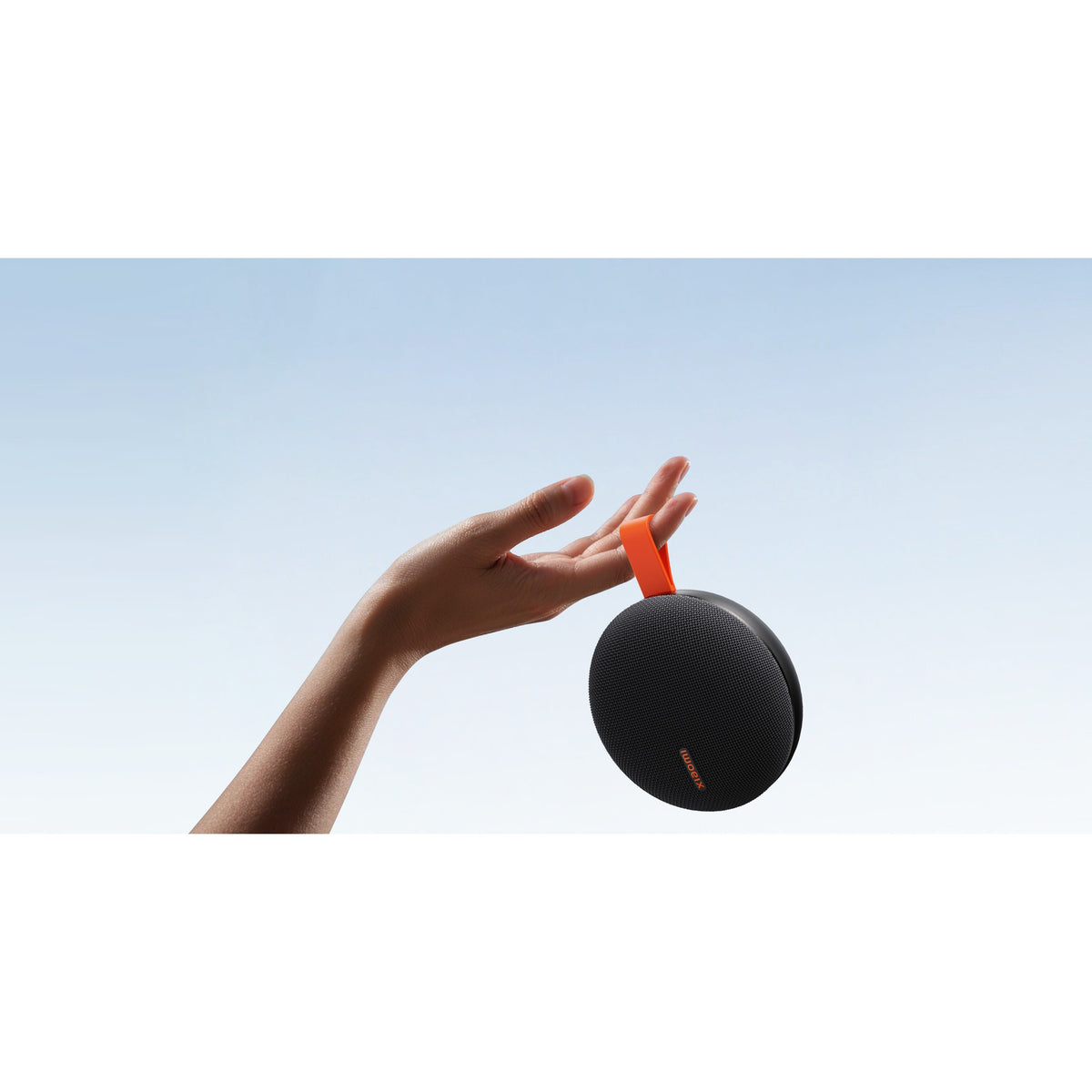 Xiaomi Bluetooth Speaker Essential in black held by its orange strap from one hand, showing how small and light the speaker is